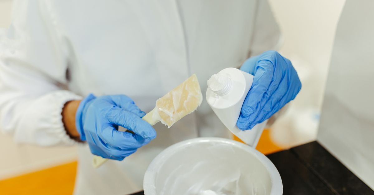 Una persona con guanti blu e camice bianco applica del silicone bianco con una spatola, tenendo in mano un tubetto di sigillante sopra un contenitore.