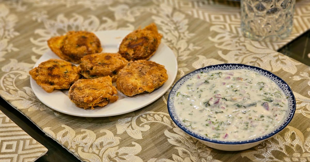 Piatto di frittelle fritte accanto a una ciotola di raita allo yogurt con cetriolo e spezie tritate.