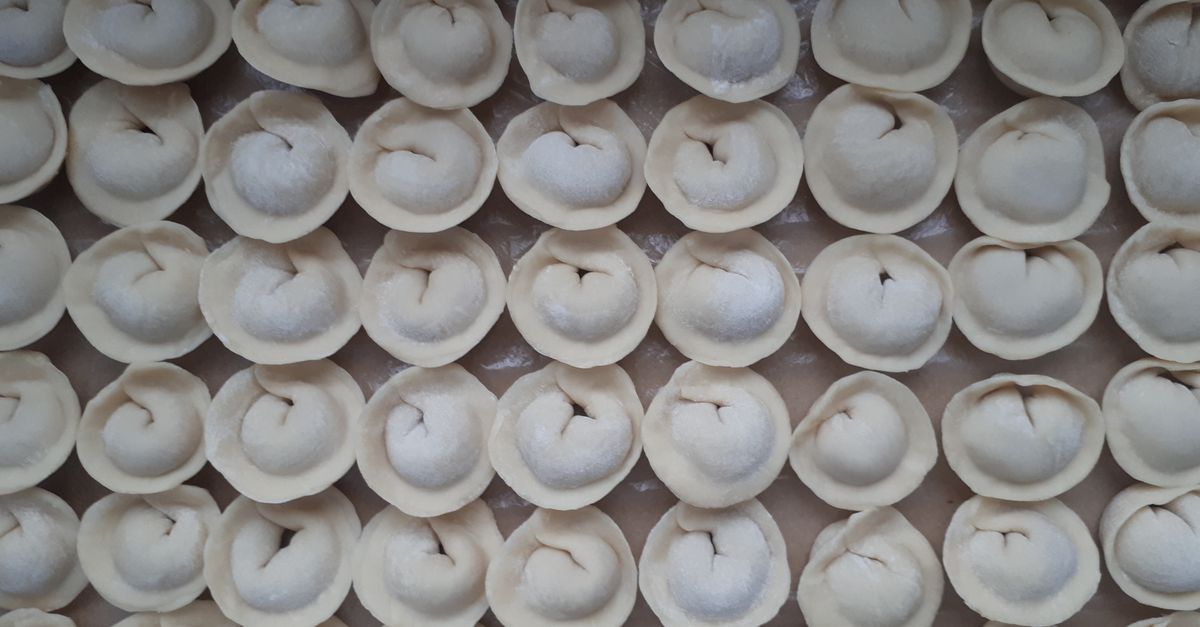 File ordinate di ravioli crudi disposti separatamente su una teglia infarinata, pronti per il congelamento.
