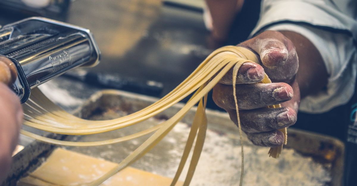 Mani infarinate estraggono tagliatelle fresche da una macchina per la pasta, con un vassoio infarinato sullo sfondo
