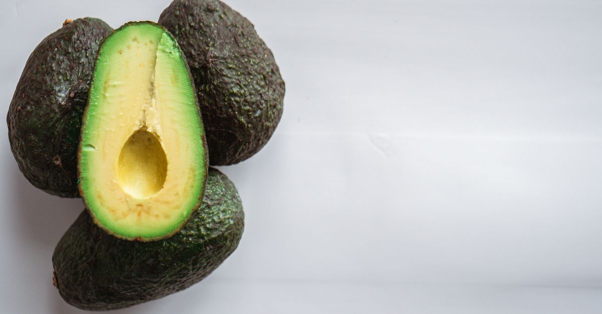 Un avocado tagliato a metà con la polpa verde brillante, appoggiato accanto ad avocado interi su sfondo bianco.