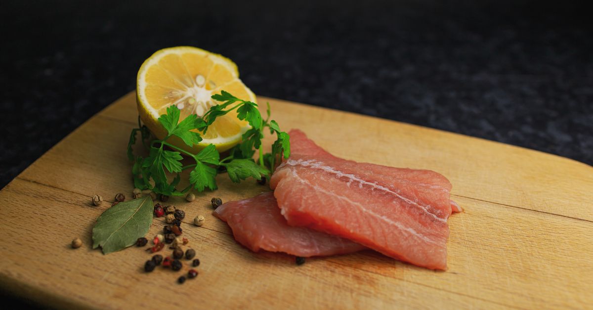 Filetti di pesce crudo su un tagliere di legno accompagnati da limone, prezzemolo, foglia di alloro e grani di pepe.