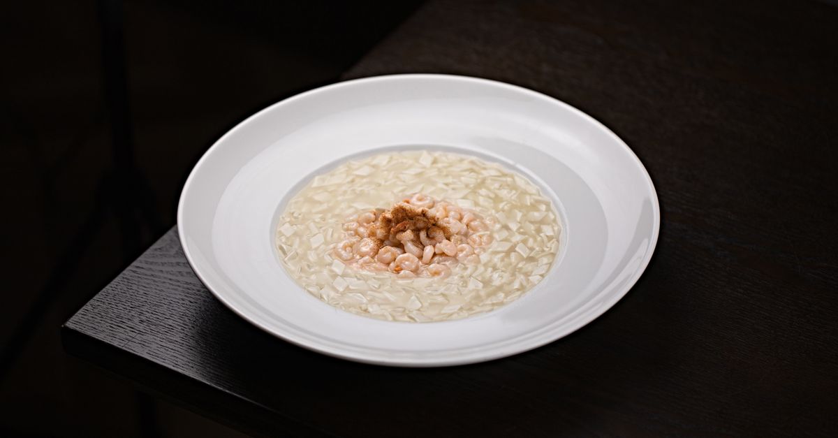 Un piatto bianco con risotto cremoso guarnito al centro con gamberetti, servito su un tavolo scuro.