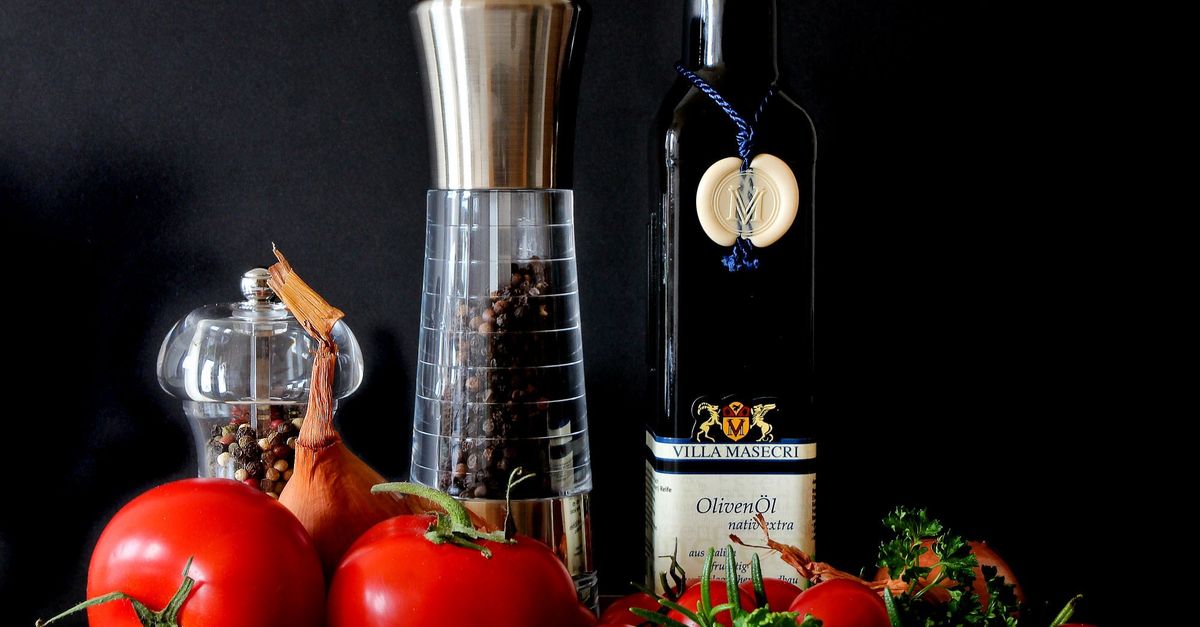 Bottiglia scura di olio extravergine con etichetta, accanto a pomodori freschi, macinapepe e cipolla su sfondo scuro