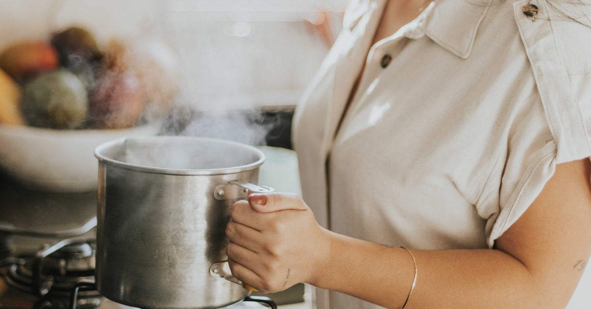 Una donna tiene con un guanto il manico di una pentola di acciaio da cui esce vapore, con acqua in ebollizione all'interno.