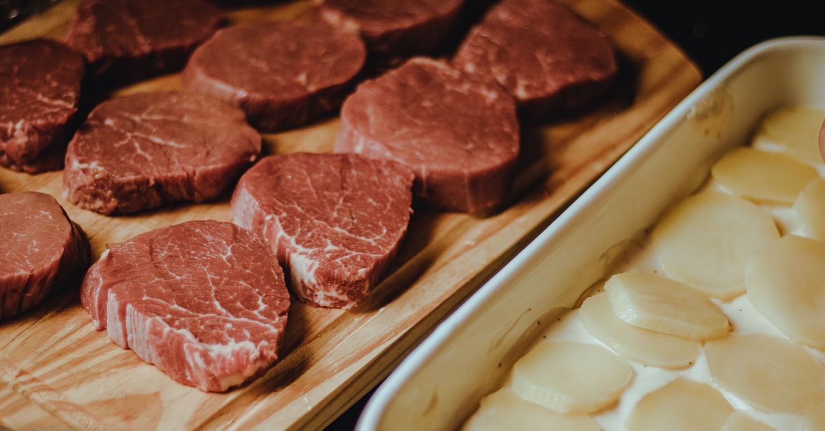 Fette di carne di manzo crudo disposte su un tagliere di legno accanto a una teglia di patate a fette.