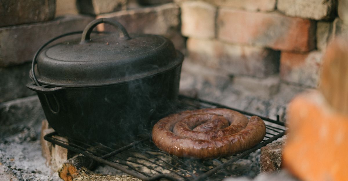 Una pentola di ghisa nera sul fuoco all'aperto, accanto a un salsicciotto a spirale che cuoce su una griglia