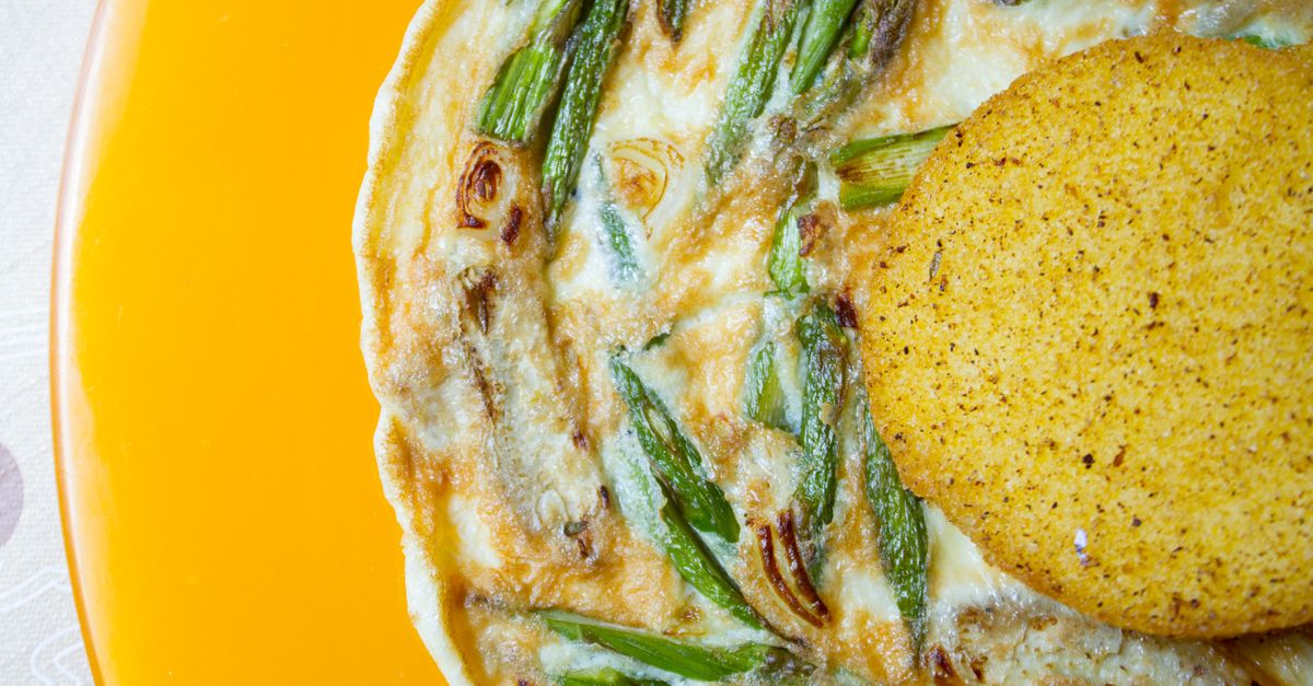 Vista dall'alto di una frittata con asparagi e cipollotti in una teglia gialla, servita accanto a una fetta di polenta grigliata.