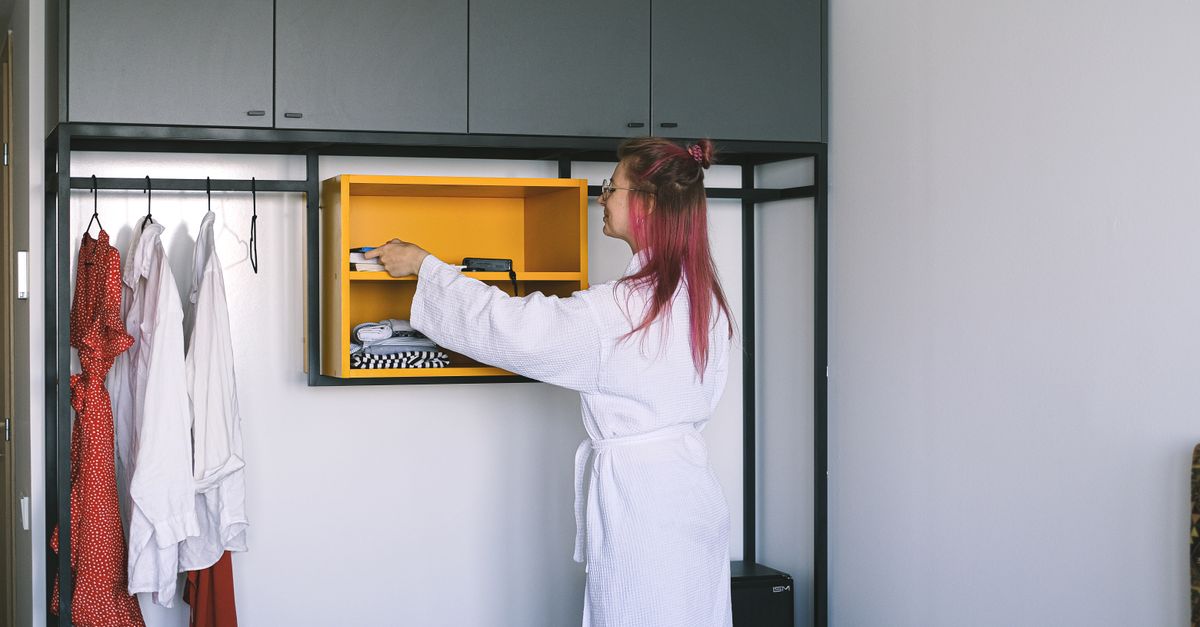 Una donna in accappatoio bianco organizza un ripiano giallo all'interno di un armadio aperto, con abiti appesi accanto.