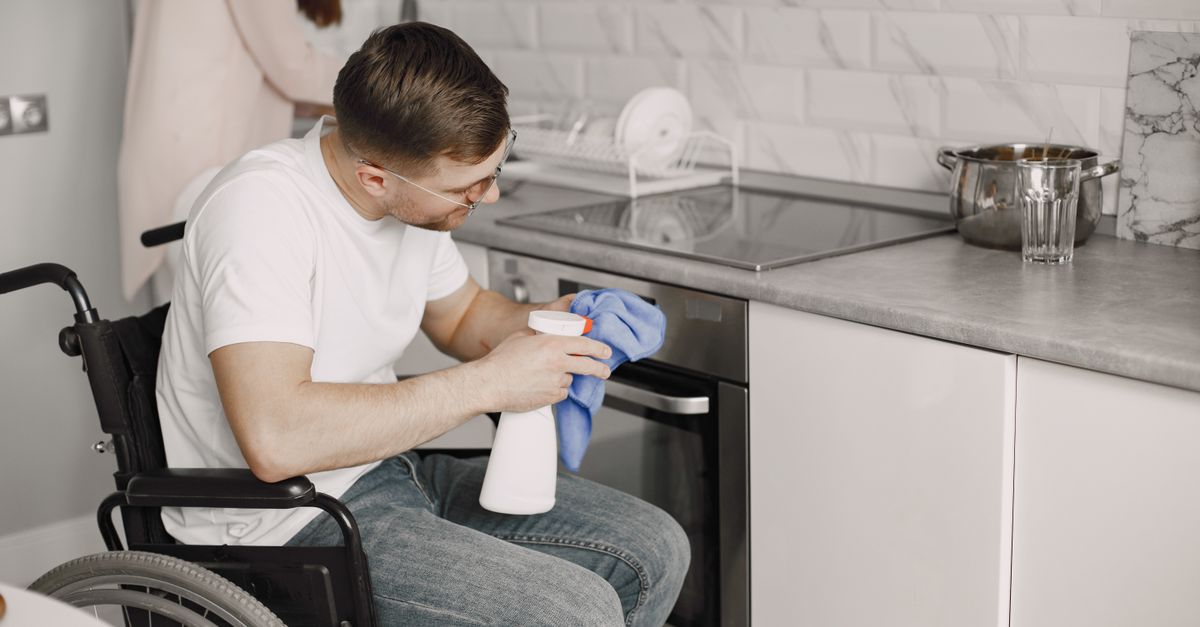 Un uomo in sedia a rotelle pulisce l'acciaio inox del forno con un panno in microfibra blu e una bottiglia spray, in una cucina moderna con lavello in acciaio inox e pentola sul piano di lavoro.