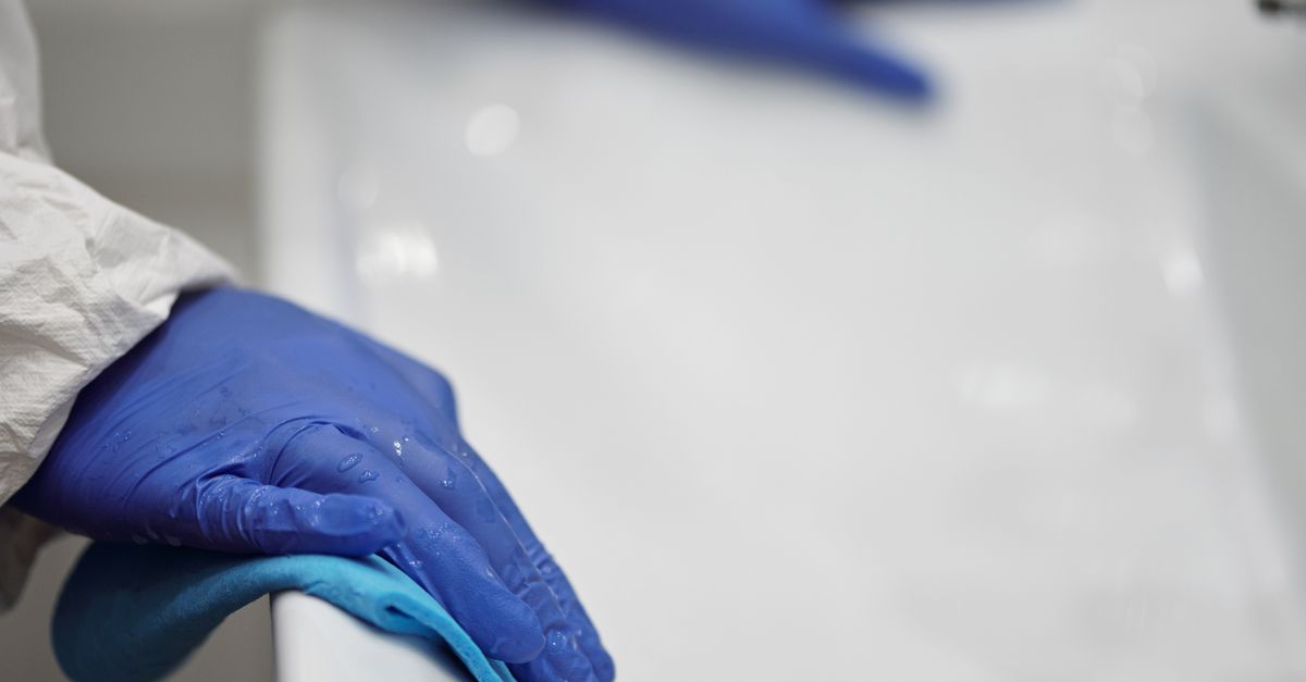 Una mano con guanto blu in lattice pulisce con un panno una superficie bianca del bagno.