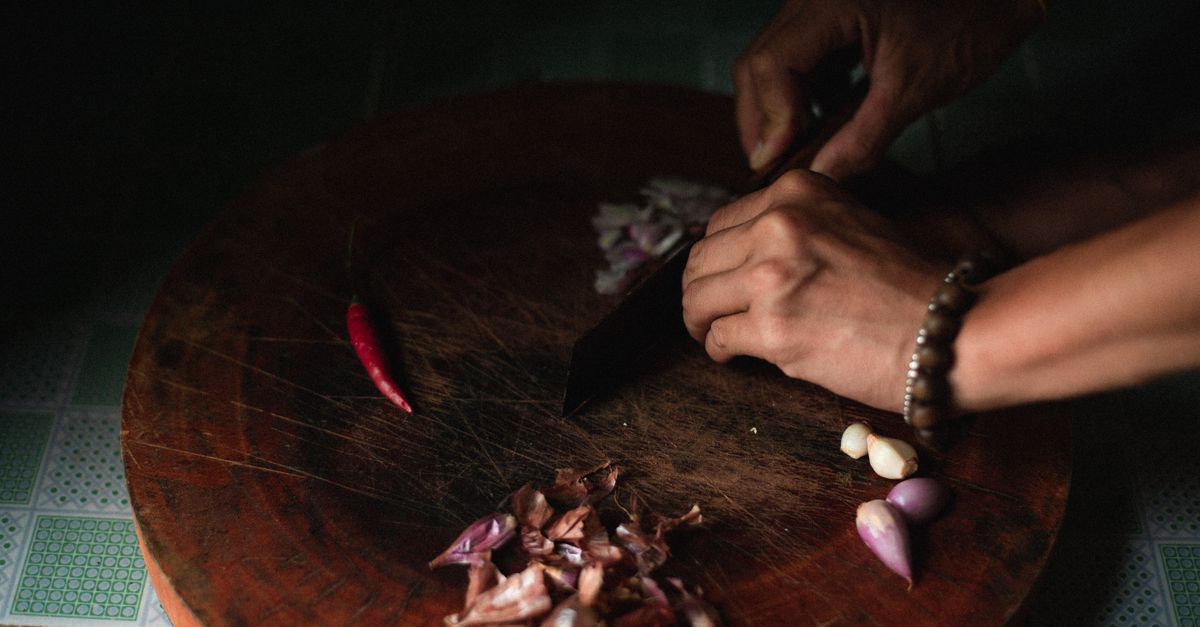 Mani che tagliano piccole cipolle rosse e aglio su un tagliere di legno scuro, con un peperoncino accanto.
