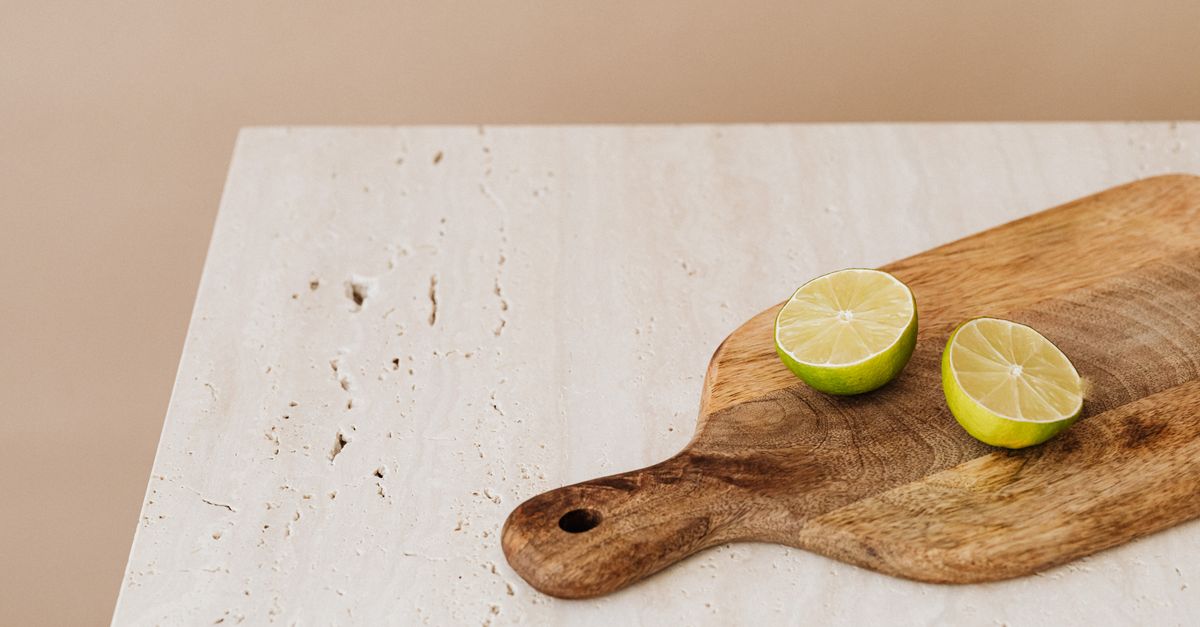 Un piccolo tagliere di legno con manico, appoggiato su un piano chiaro, con due metà di lime tagliate a fette poste sopra.