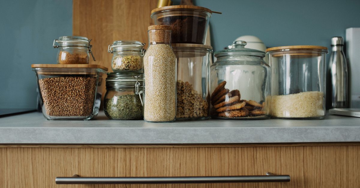 Barattoli di vetro trasparenti con coperchi ermetici, pieni di legumi, cereali e spezie, allineati su un piano cucina.