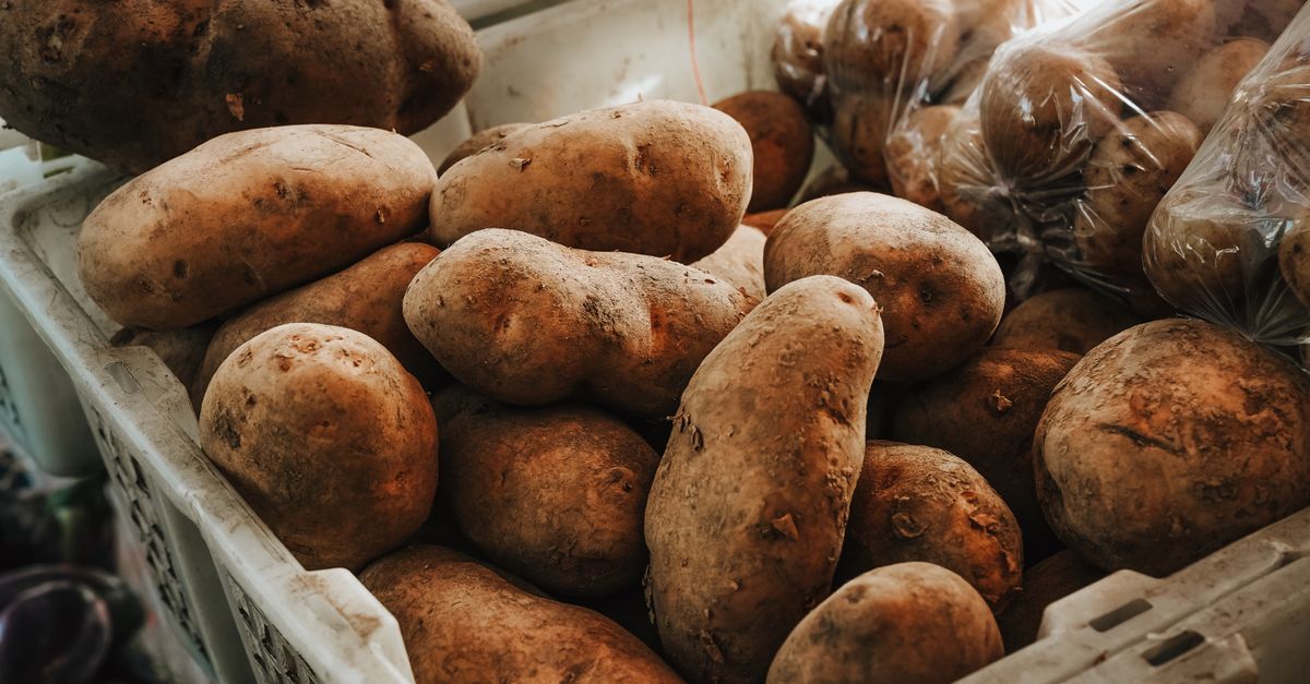 Cassetta piena di patate grezze e terrose accatastate insieme ad altre confezionate in sacchetti di plastica.