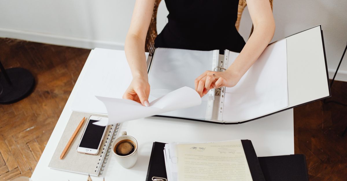 Una persona seduta a una scrivania bianca sfoglia documenti organizzati in un raccoglitore ad anelli, con quaderno, penna e tazzina di caffè accanto.