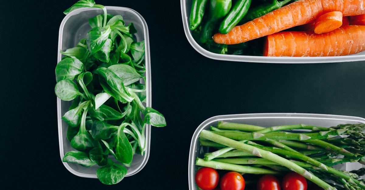 Vista dall'alto di contenitori trasparenti con verdure fresche separate, tra cui insalatina, carote, fagiolini, asparagi e pomodorini.