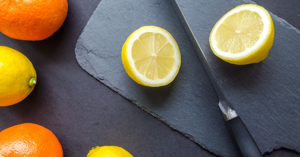 Limoni tagliati a metà accanto ad altri agrumi interi su un tagliere scuro, con un coltello appoggiato accanto.