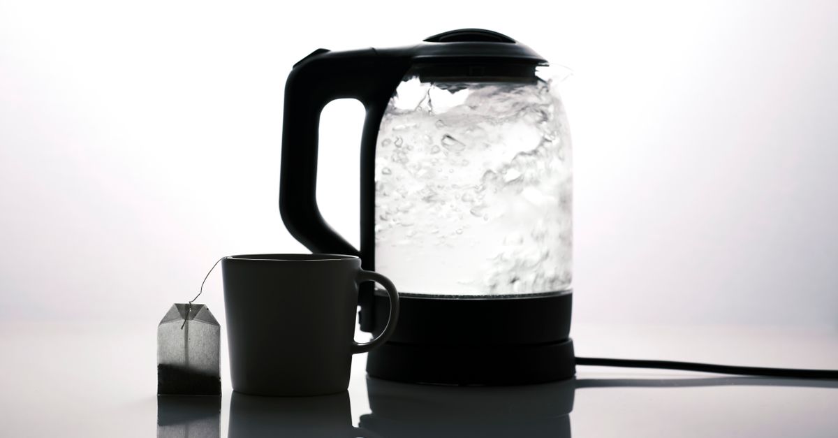 Un bollitore elettrico trasparente con acqua bollente in ebollizione, accanto a una tazza bianca con una bustina di tè.