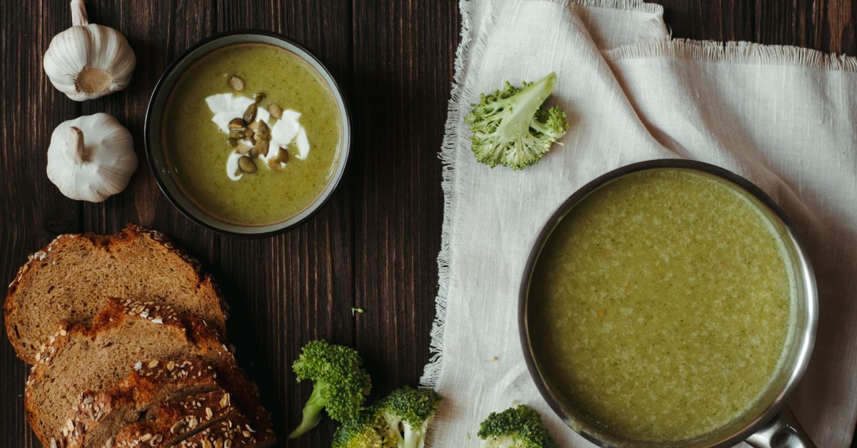 Ciotola di crema di broccoli con panna e semi, servita accanto a fette di pane integrale e teste d'aglio su un tavolo di legno scuro