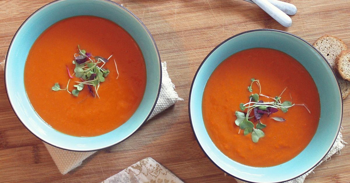 Due ciotole di crema di pomodoro arancione servite in scodelle blu con guarnizione di germogli, accanto a fette di pane su un tavolo di legno.