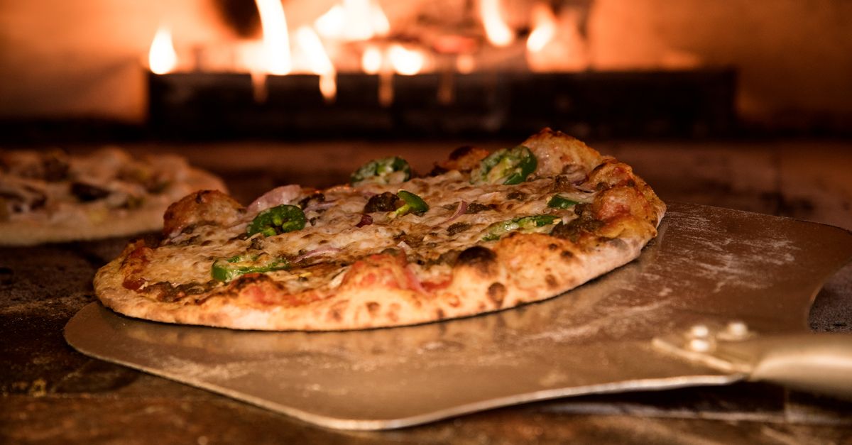 Una fetta di pizza appena tolta dal forno a legna, su una pala metallica, con le fiamme sullo sfondo.