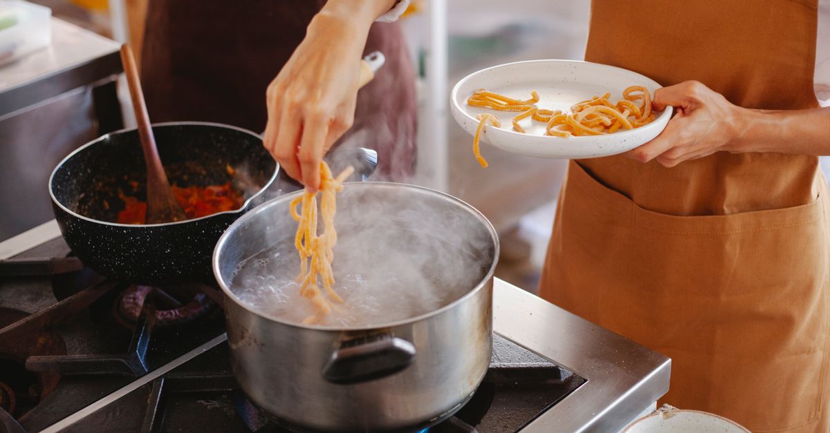 Una persona solleva spaghetti dalla pentola di acqua bollente mentre tiene un piatto vuoto, con una padella di condimento sullo sfondo.