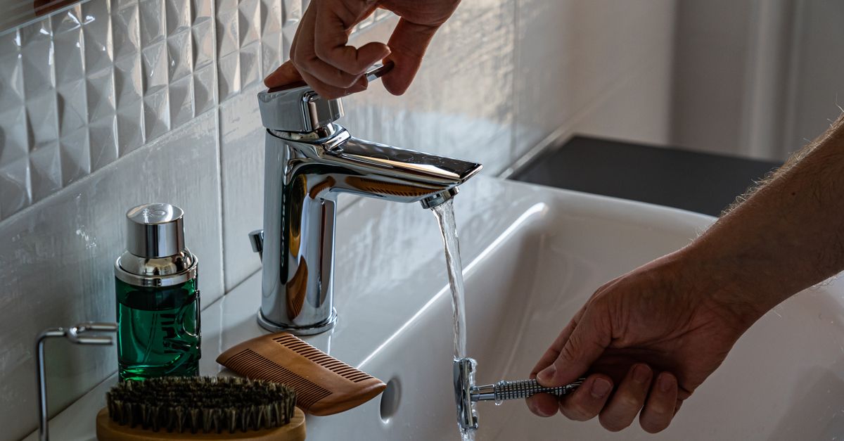 Mani di una persona che regolano il rubinetto cromato di un lavandino da bagno, con un pettine e un flacone verde appoggiati sul bordo.