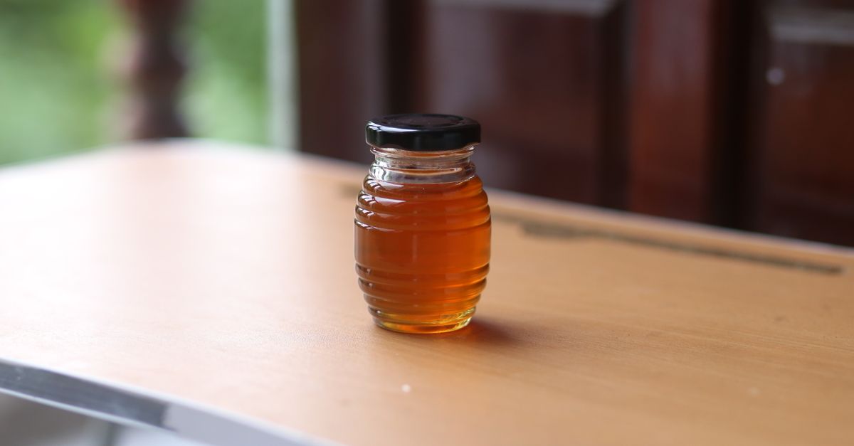 Un barattolo di vetro con miele liquido e coperchio nero appoggiato su un tavolo di legno chiaro.