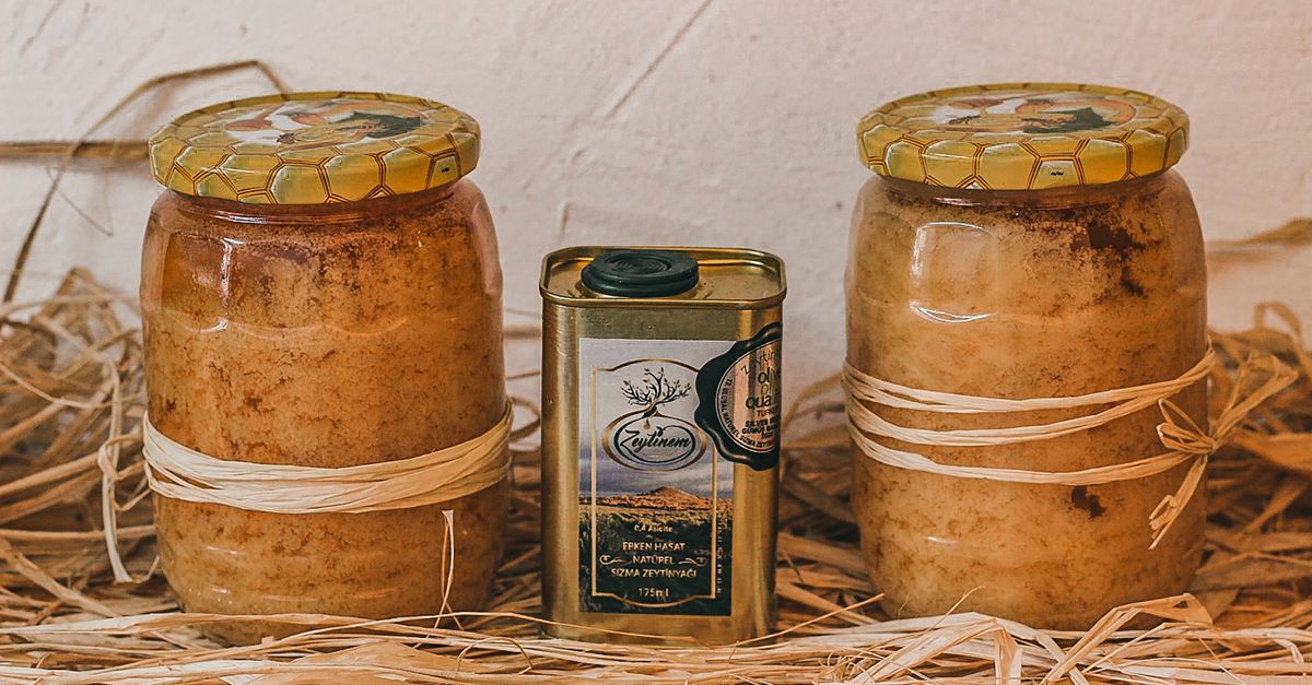 Due grandi vasi di vetro pieni di miele cristallizzato con coperchi decorati, affiancati da una latta di olio, su un letto di paglia.
