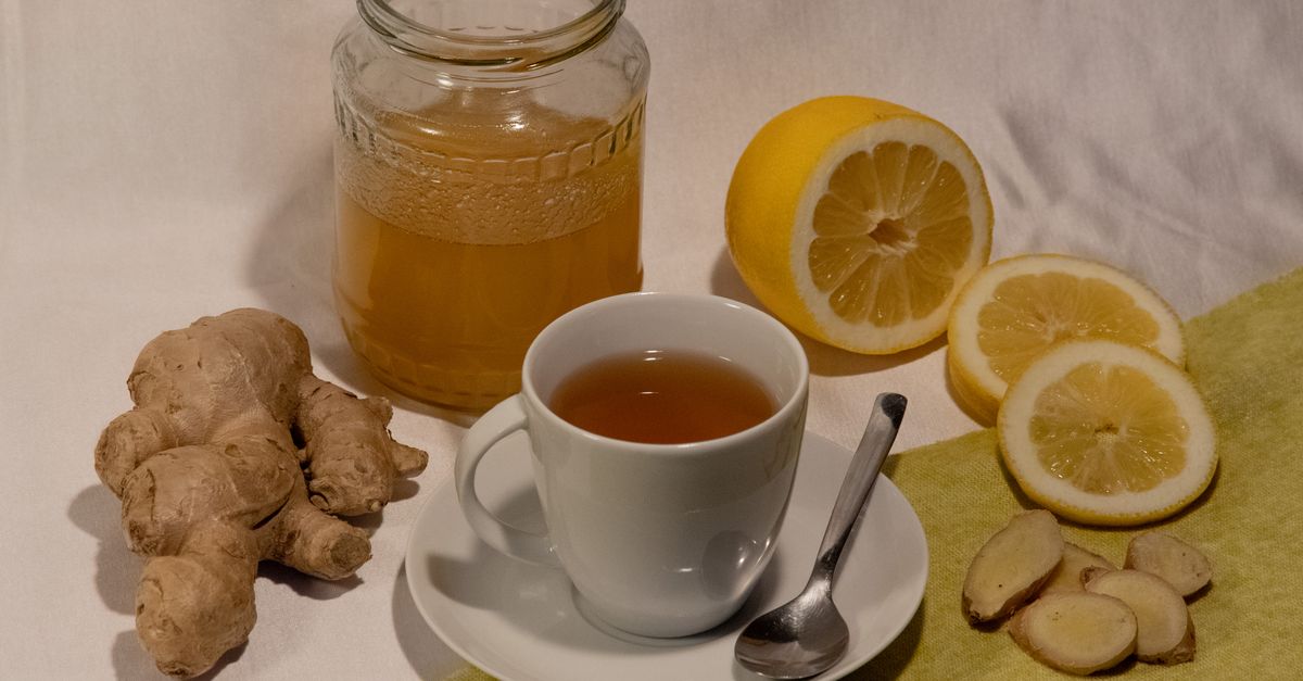 Radice di zenzero fresco accanto a una tazza di tè, miele in barattolo e limoni tagliati su un tavolo.