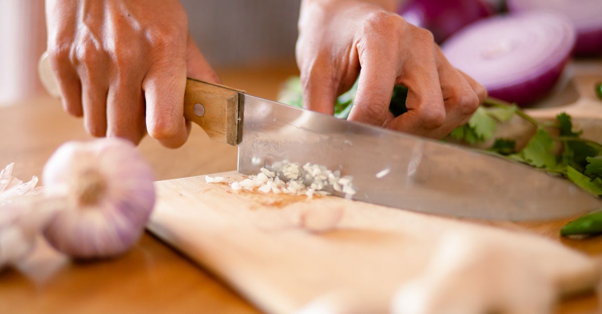 Mani che tritano aglio con un coltello da chef su un tagliere di legno, con cipolla rossa e coriandolo sullo sfondo.