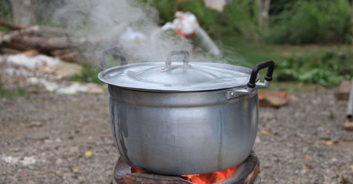 Una pentola in metallo con coperchio fuma sopra un fuoco all'aperto, con brace rossa visibile sotto.