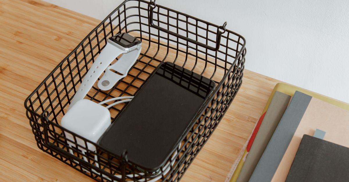 Un cestino metallico su una scrivania di legno contiene uno smartwatch e uno smartphone con il relativo cavo di ricarica ordinato.
