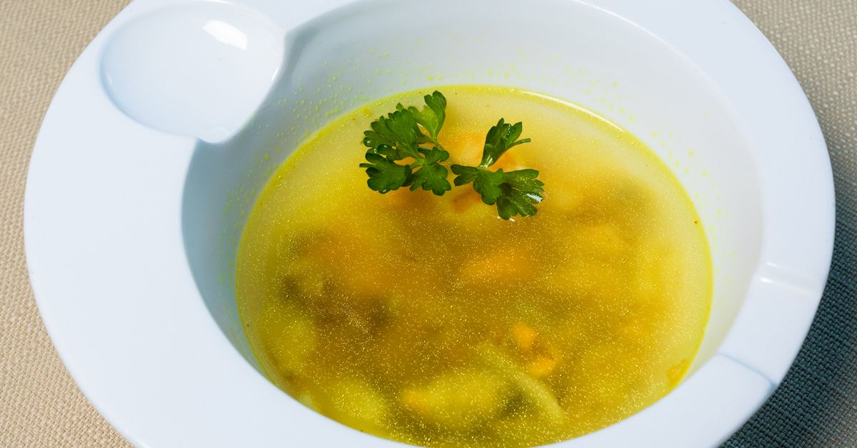 Una ciotola bianca con brodo vegetale limpido e dorato, con pezzetti di verdura e una foglia di prezzemolo fresco al centro.