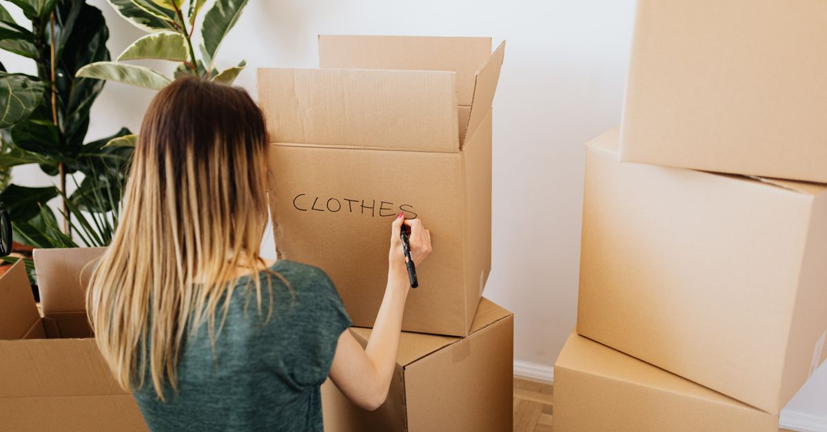 Una donna scrive a mano la parola "CLOTHES" su una scatola di cartone circondata da altre scatole per il trasloco.