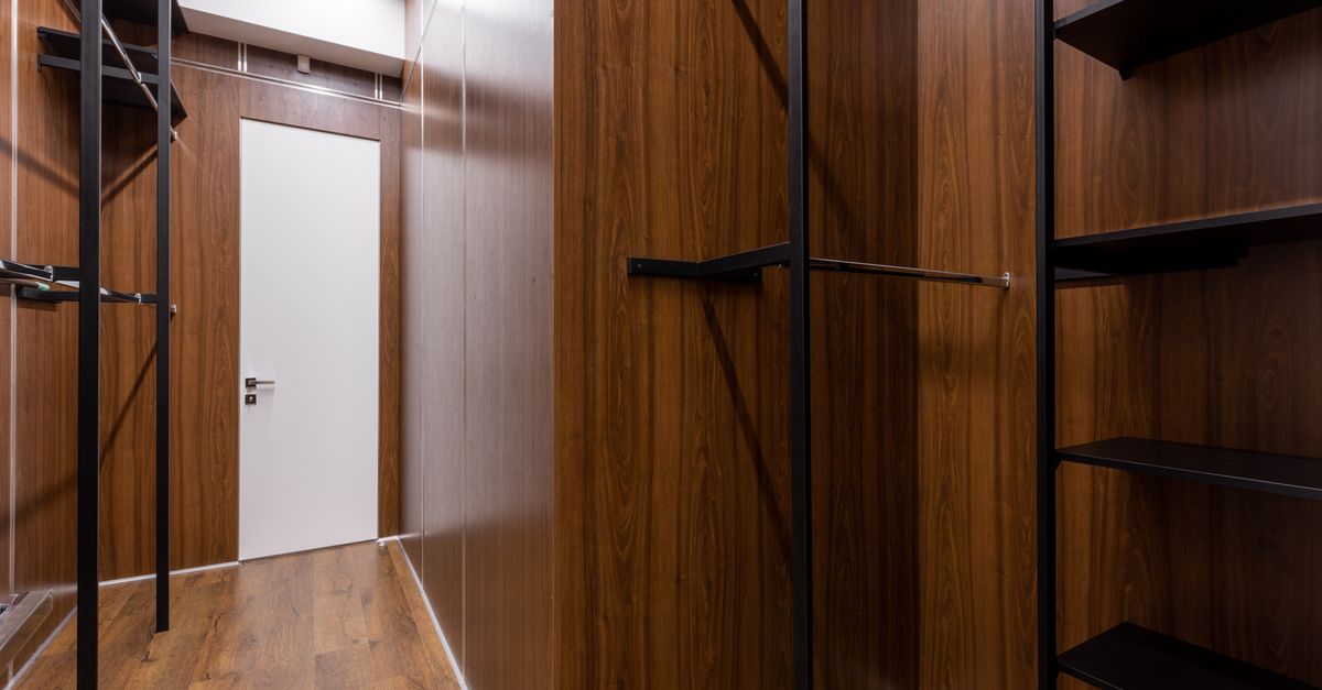 Cabina armadio con pareti e mensole in legno scuro, porta bianca sullo sfondo e pavimento in parquet.