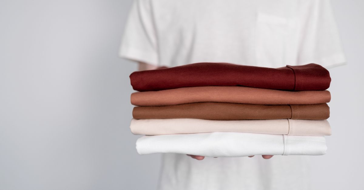 Una persona tiene tra le mani una pila di magliette piegate in diverse tonalità, dal bordeaux al bianco.