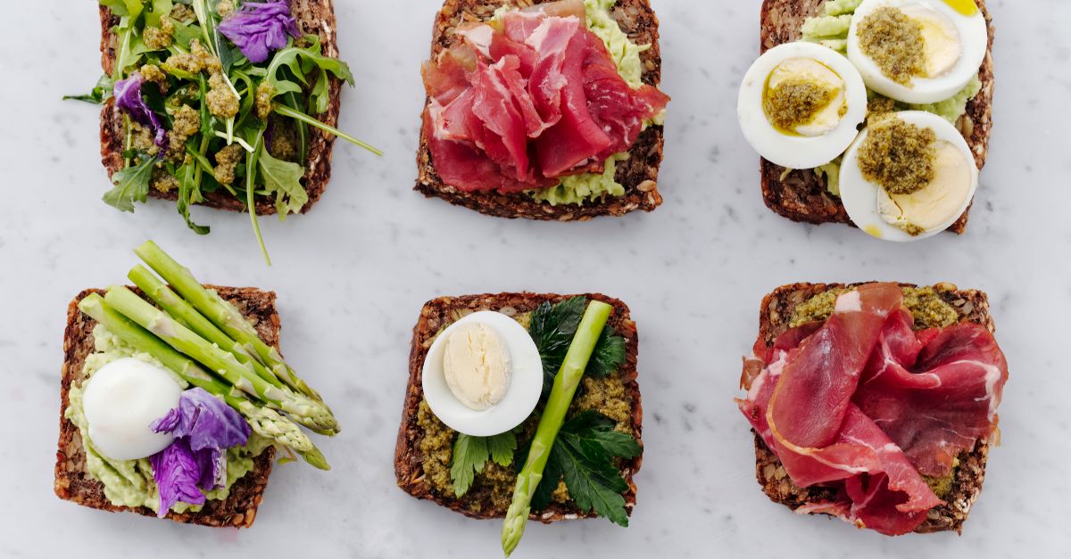 Toast di pane integrale con avocado, asparagi, uova sode affettate e prosciutto, disposti su un tavolo chiaro in stile marmo.
