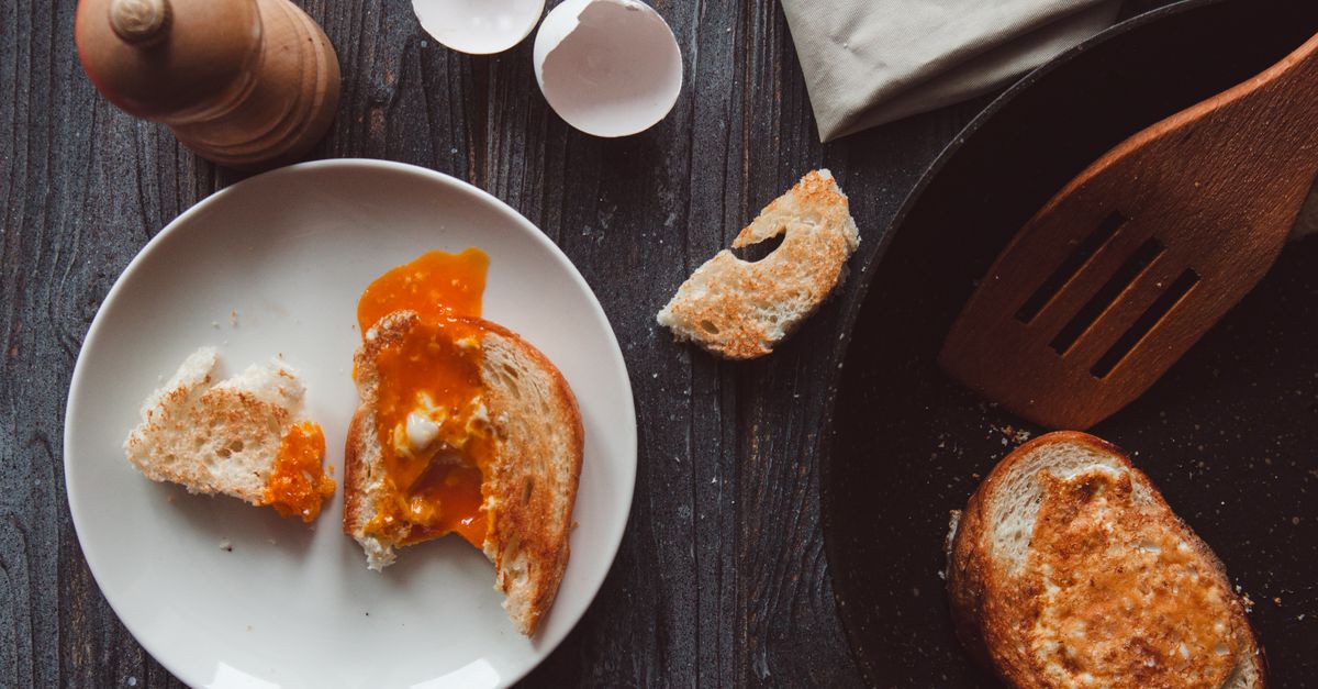 Toast con uovo morbido e tuorlo che cola, servito su un piatto accanto a un guscio d'uovo e una padella.